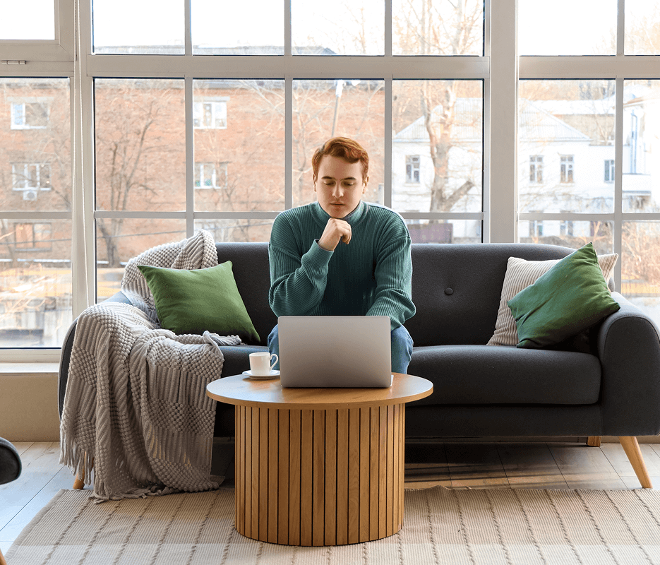 A man with short, red hair is sitting on a couch and looking at a laptop on a coffee table. He is surrounded by icons of a lock, a lightning bolt, and a shield with a checkmark in the center.
