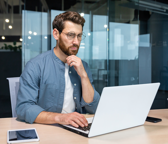 A man with a well-trimmed beard is looking at a laptop curiously.