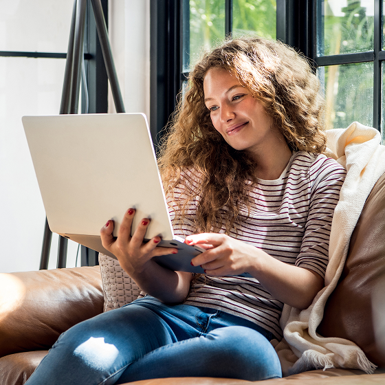 A woman with long, curly hair is sitting in a sunny alcove holding a laptop and smiling at the way Tweaknews’ security solutions are protecting her from threats online.