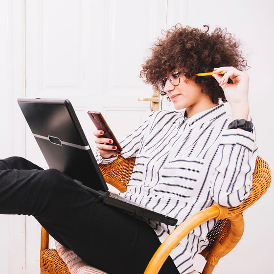 A woman with wild hair is leaning back in a wicker chair with a laptop on her lap. She is looking on her phone for good newsgroups to search for and absently hitting her head with a pencil eraser.
