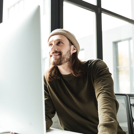 A man with long brown hair and a tan beanie cap is excitedly looking at newsgroups on his desktop monitor.