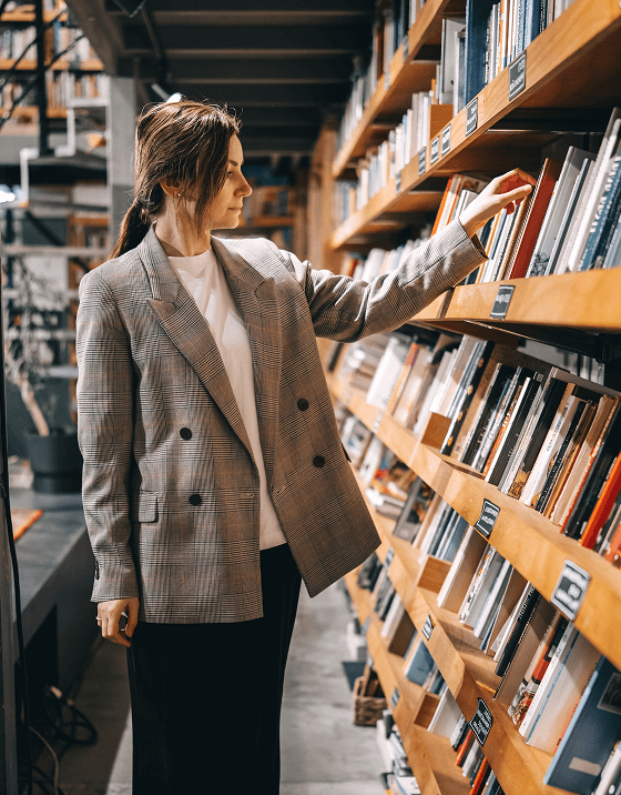 A woman in a stylish tweed sport coat is browsing books in a large library.