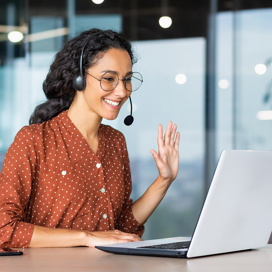 A woman with large, wire-rimmed glasses is wearing a headset and smiling at a computer, helping a Tweaknews subscriber.