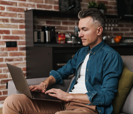 A man with black hair going grey at the temples and wearing a denim button-down shirt is typing on a laptop.
