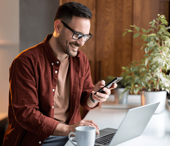 A man with short, black hair and glasses is smiling while reading something on his phone. With his other hand, he is connecting to a VPN through Tweaknews on his laptop, since it’s so easy he can do that with a single click.