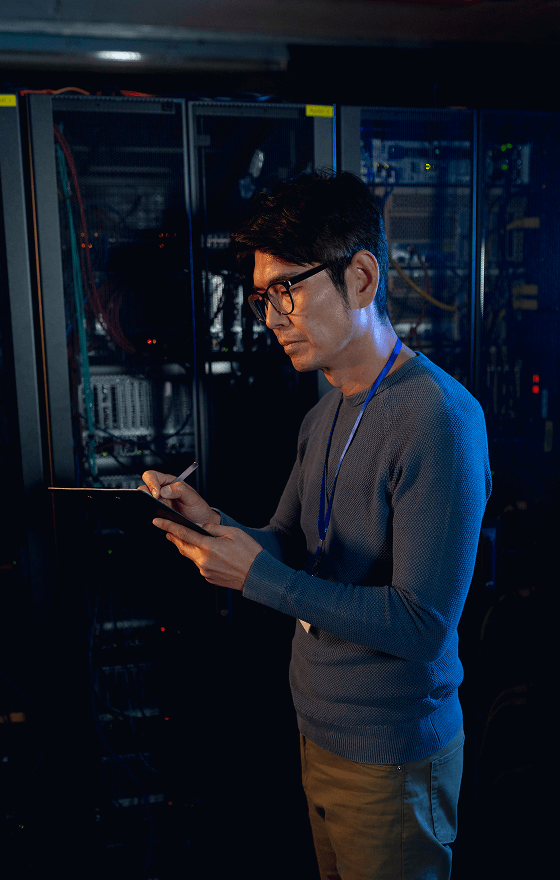 A serious man in glasses is standing in a Usenet server farm and taking notes on a tablet.