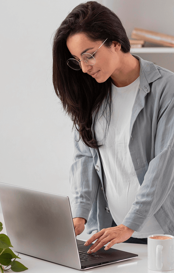 A woman with round, wire-rimmed glasses is standing in front of a desk and typing on a laptop.