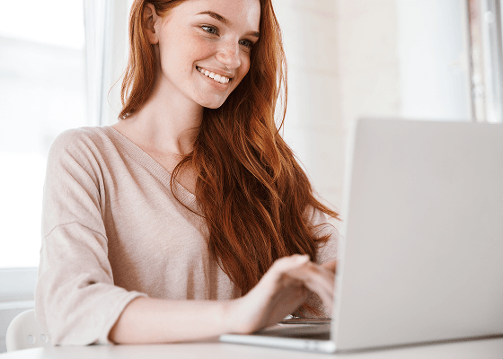 A woman with long, red hair is on a laptop, smiling because she just signed up for Tweaknews and was able to almost immediately start enjoying Usenet.