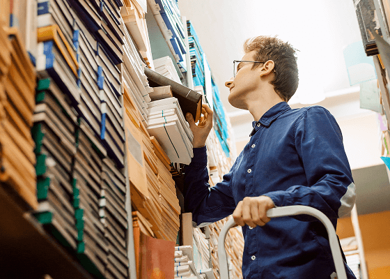 A man is standing on a stepstool, grabbing a book from a large stack of them.