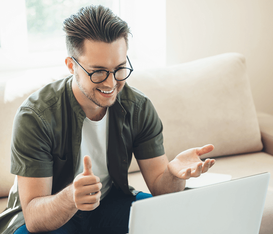 A man with slicked back hair and round, black glasses is giving a thumbs up to his laptop.