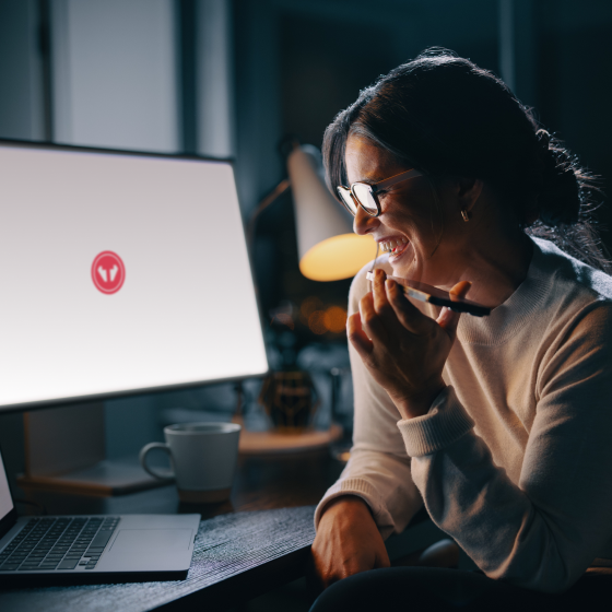 Une femme tient son téléphone portable comme si elle parlait en haut-parleur. En arrière-plan se trouve un écran d’ordinateur avec le logo Tweaknews.