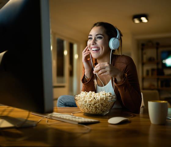 Une femme est assise devant un ordinateur de bureau, écoute quelque chose avec son casque et mange du pop-corn en riant.